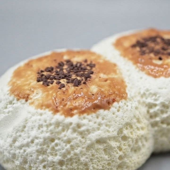 Two round white meringue-based pastries with golden-brown centers and dark chocolate sprinkles on top, photographed on a smooth gray surface.