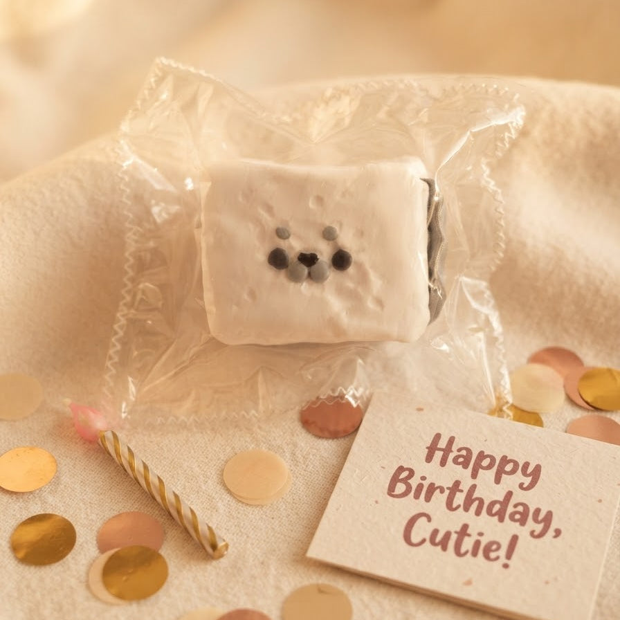 A cute, square-shaped white foam squishy toy with a bear face, wrapped in clear cellophane packaging. It is styled on a soft beige fabric with gold and pink confetti, a small striped candle, and a card that reads 'Happy Birthday, Cutie!'.