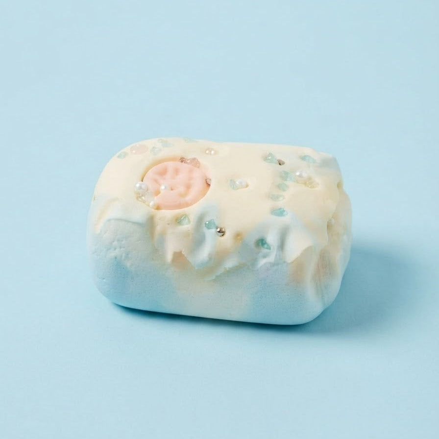 A single bath bomb with a rounded rectangular shape, featuring a pale cream top with white marbling, light blue speckles, and a small pink circular detail, set against a solid light blue background.