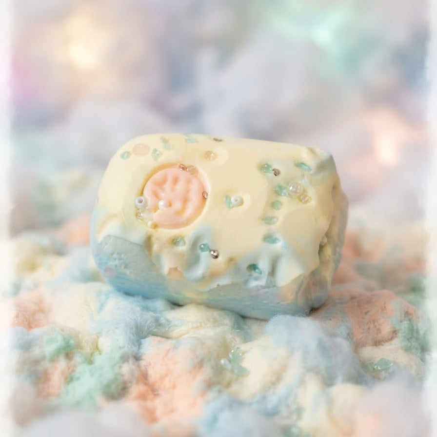 A pastel-colored bath bomb with a pale yellow top, light blue sides, and tiny green and silver sprinkles, resting on a bed of fluffy pastel cotton candy.