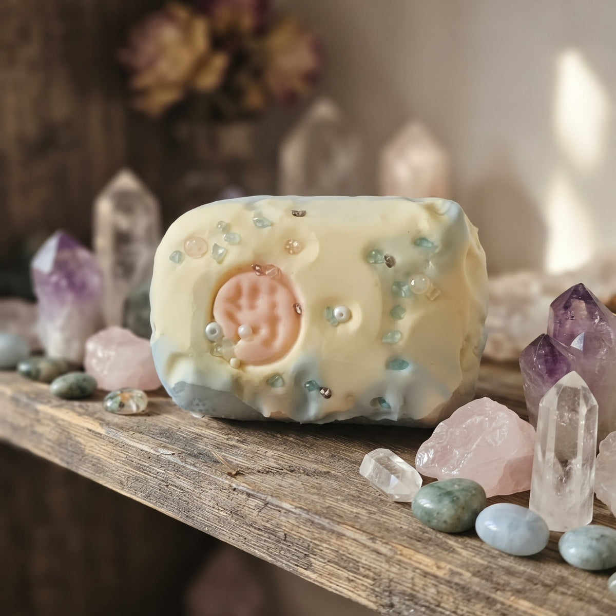 A handmade soap bar with a pale yellow and white marbled finish, decorated with tiny blue and green glitter flecks and a central pink circular motif, displayed on a rustic wooden shelf surrounded by crystals.