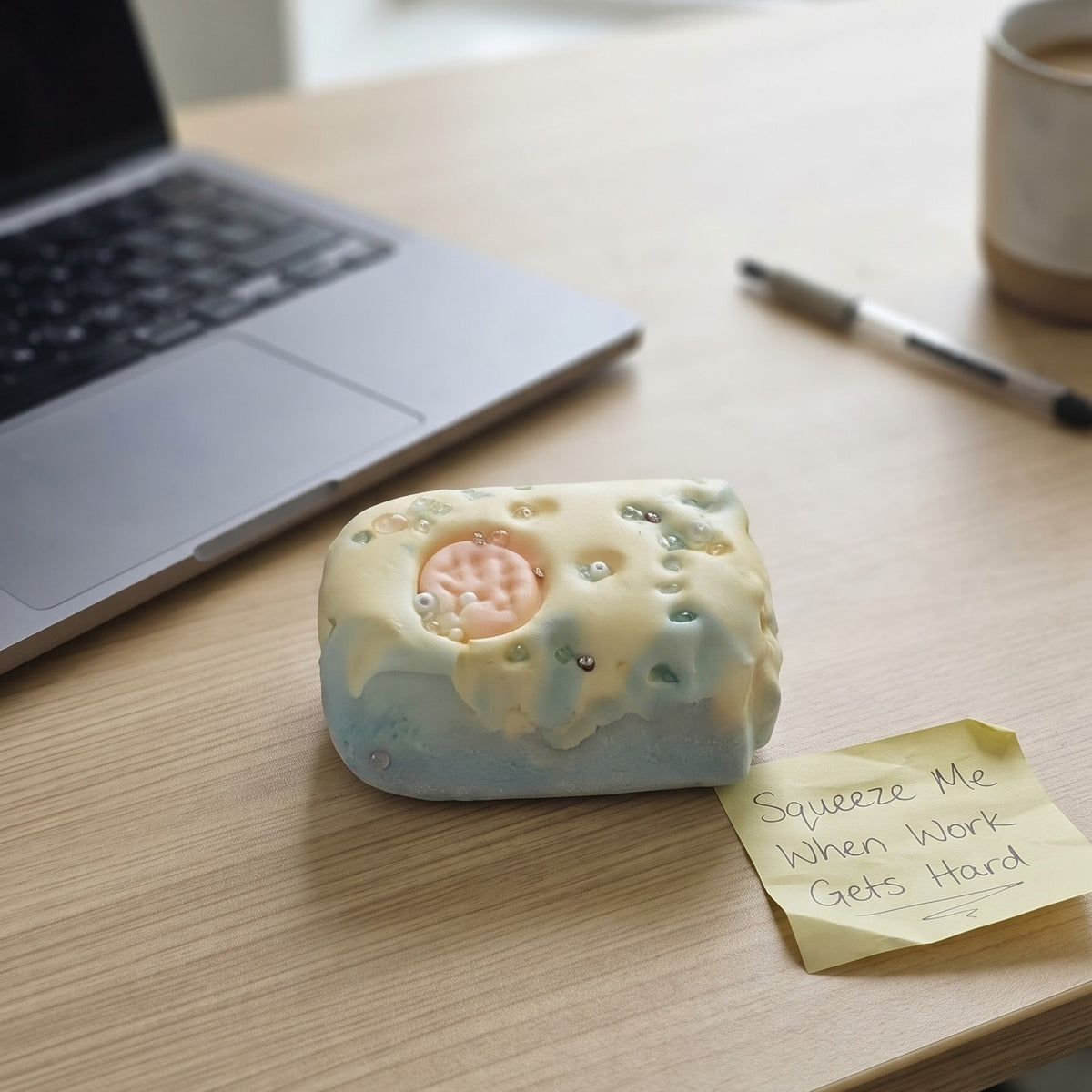 A small rectangular novelty object with a pale blue base and cream-colored top sits on a wooden desk next to a sticky note that reads 'Squeeze Me When Work Gets Hard.' A laptop, pen, and mug are blurred in the background near a window.