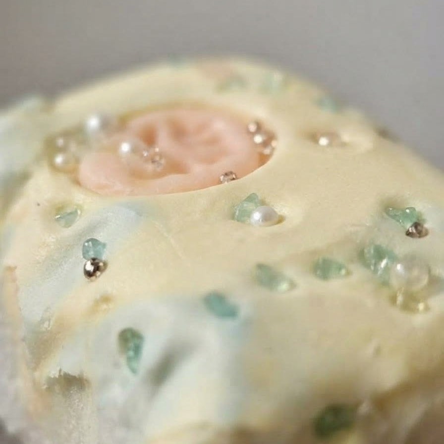 A close-up of a pale yellow and white textured item, possibly a decorative soap, candle, or cosmetic bar, featuring a central peach-pink circular inset and scattered clear and green rhinestone-like embellishments.