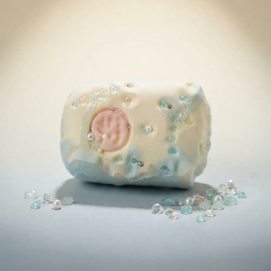 A handmade soap bar with a textured, organic shape, decorated with light blue and white marbling, small clear rhinestones, and a central pale pink circular motif. The soap is displayed on a neutral surface with scattered decorative rhinestones in the foreground under a warm spotlight.