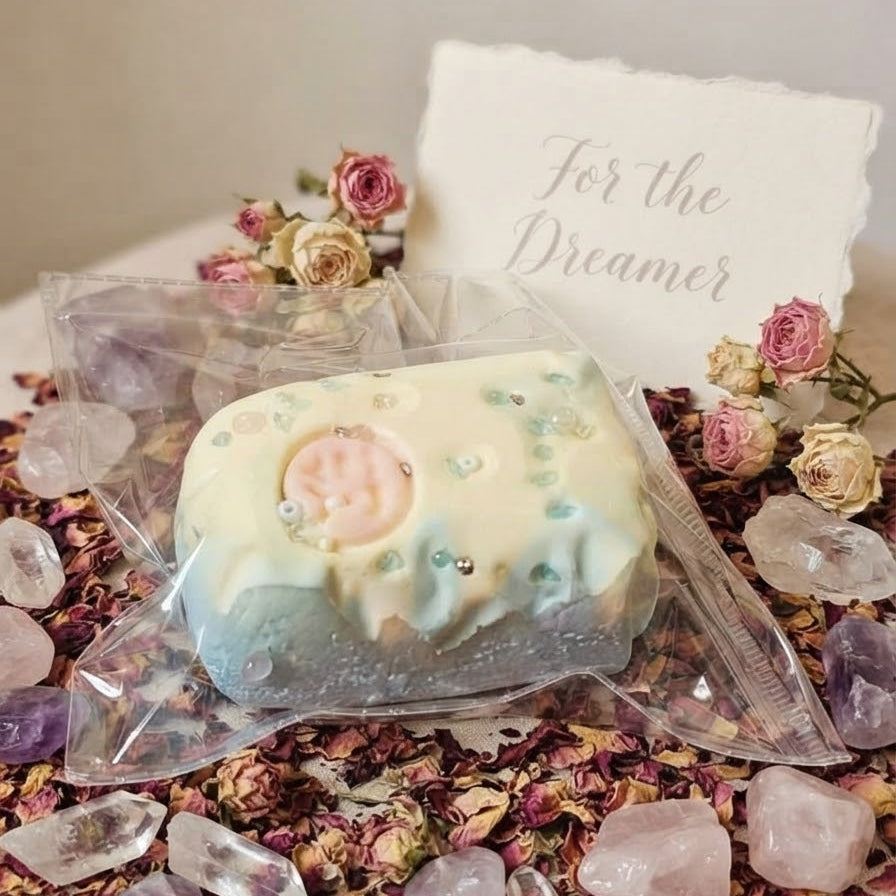 A packaged handmade bath bomb or soap bar wrapped in clear plastic, placed on a surface surrounded by dried rose petals, crystals, and small flowers. A small card behind it reads 'For the Dreamer'.