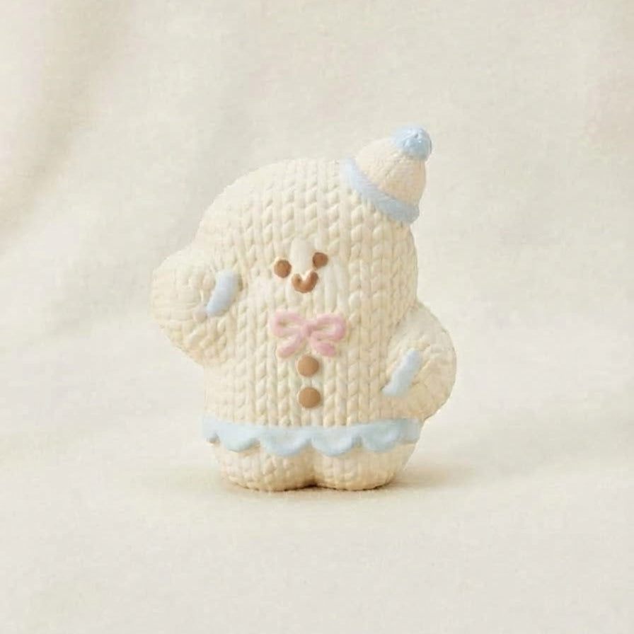 A small knitted bear figurine in cream with light blue and pink accents, standing against a soft cream fabric background.