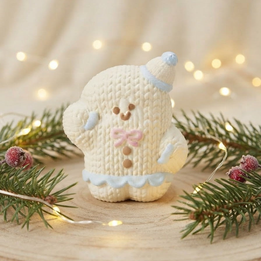 A knitted gingerbread person ornament or figurine in cream with light blue trim and a pink bow, displayed on a wooden surface with evergreen branches and warm fairy lights in the background.