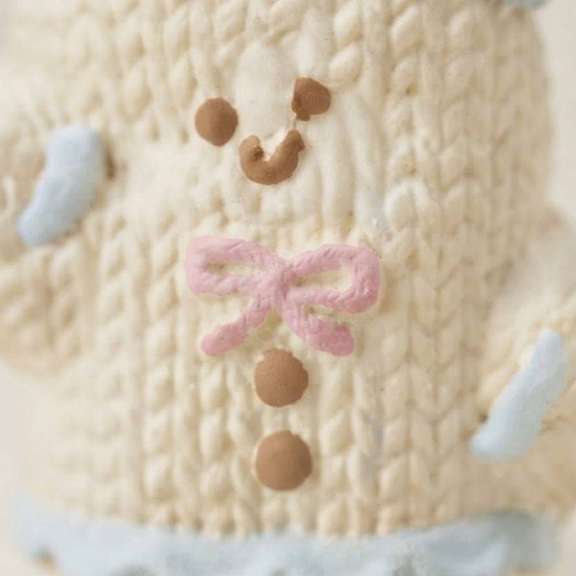 Close-up of a cream-colored knitted plush toy with a light blue hat, light blue cuffs, a pink bow, and brown embroidered facial features.