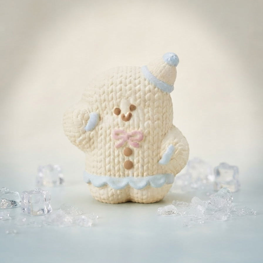 A small knitted bear figurine in cream with light blue and pink accents, standing on a pale blue surface with scattered clear ice cubes around it.