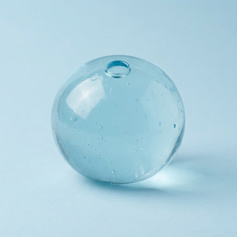 A light blue transparent glass bead with a small hole at the top, shown on a solid light blue background.