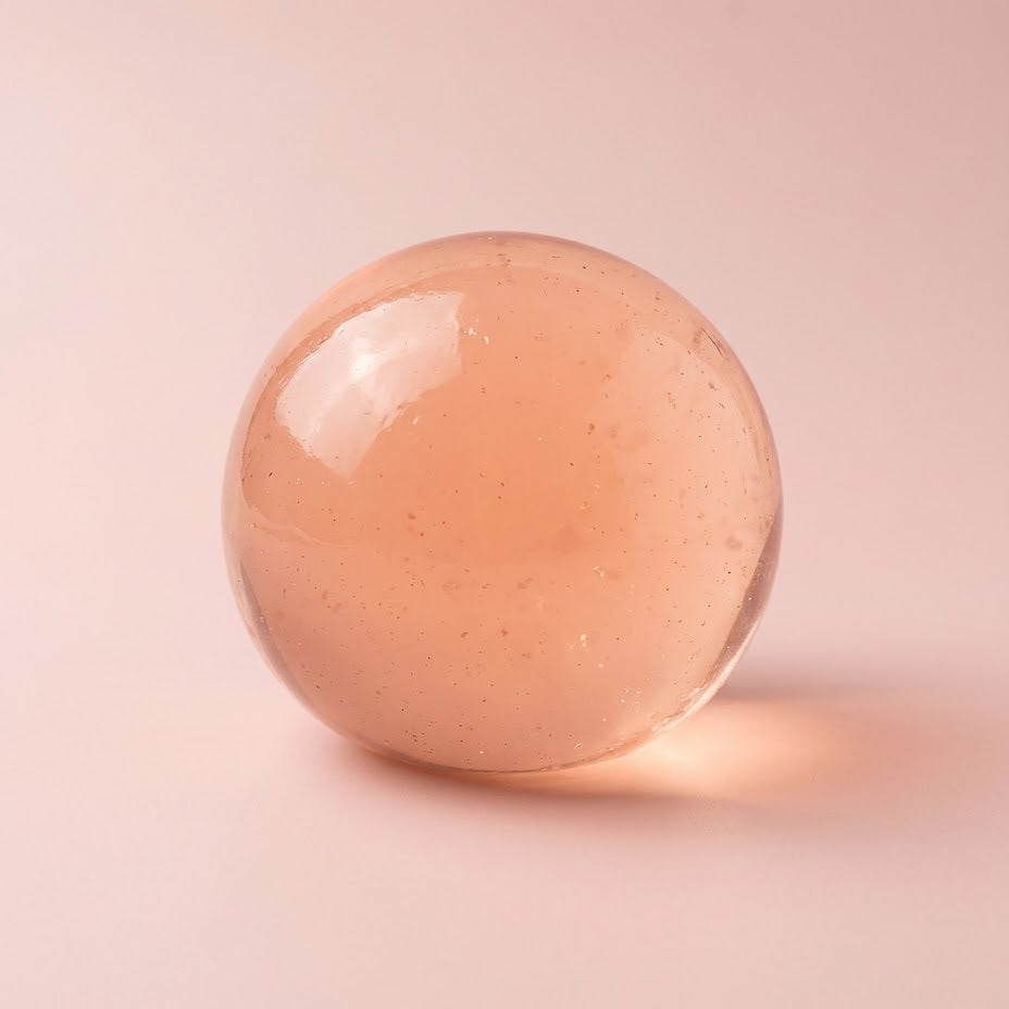 A smooth, translucent peach-colored glass sphere with tiny suspended specks, resting on a solid pale pink background.