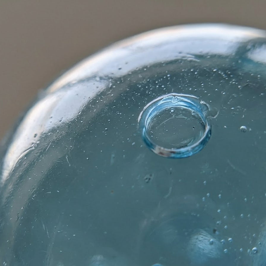 Close-up of a translucent light blue glass container or bottle with a small circular opening and visible condensation droplets, against a soft warm gray background.