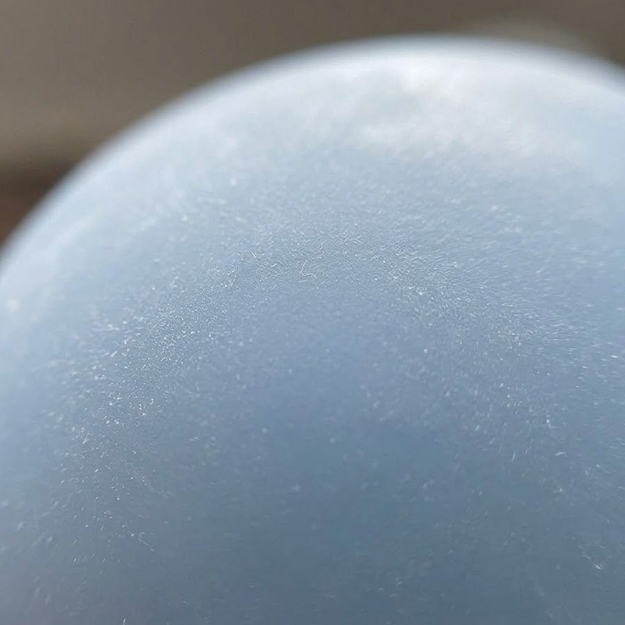 A close-up of a smooth, matte light blue sphere, possibly a decorative stone, marble, or glass orb, with a soft-focus neutral background.