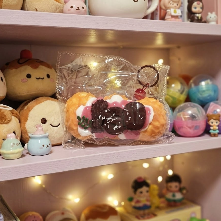 A cute, food-themed squishy toy shaped like a pastry with chocolate toppings, packaged in a clear plastic bag and displayed on a pink shelf among other plush toys and pastel items.