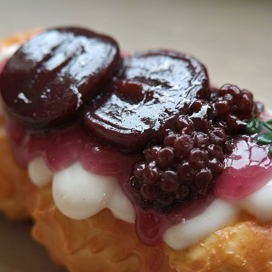 A close-up of a pastry topped with glossy dark berry slices, a berry compote, white cream, and a small green leaf garnish on a neutral background.