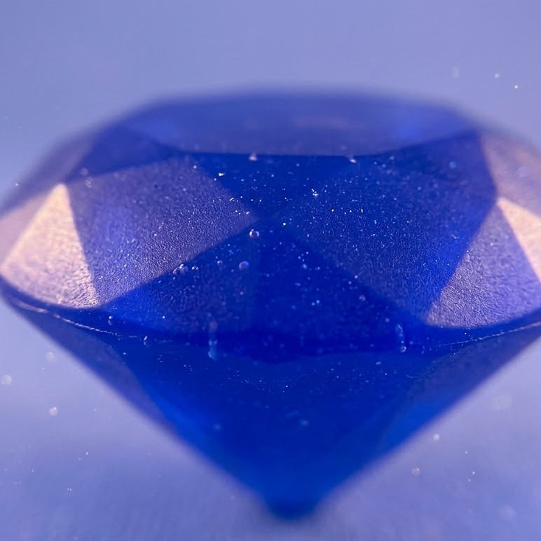 A faceted blue gemstone or decorative crystal shown close-up against a soft blue background with subtle light specks.