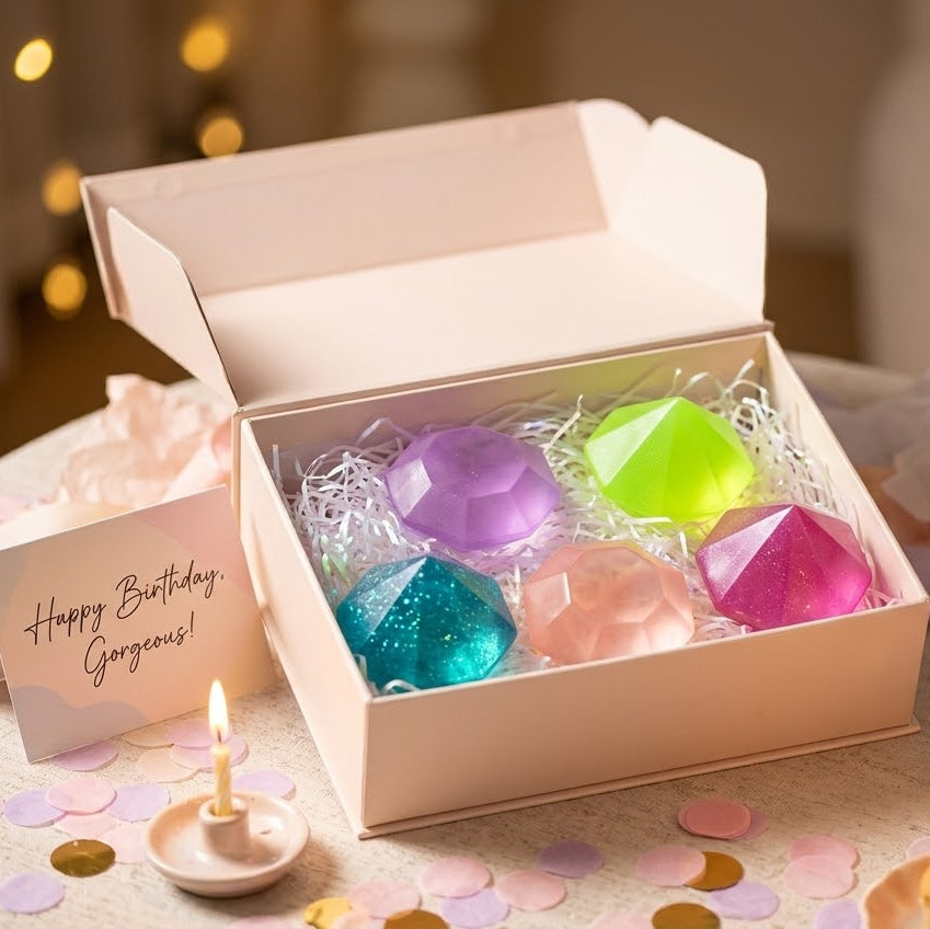 A gift box containing six faceted gem-shaped bath bombs in pastel and neon colors, displayed on a decorated birthday table with candles, confetti, and a 'Happy Birthday Gorgeous!' card.
