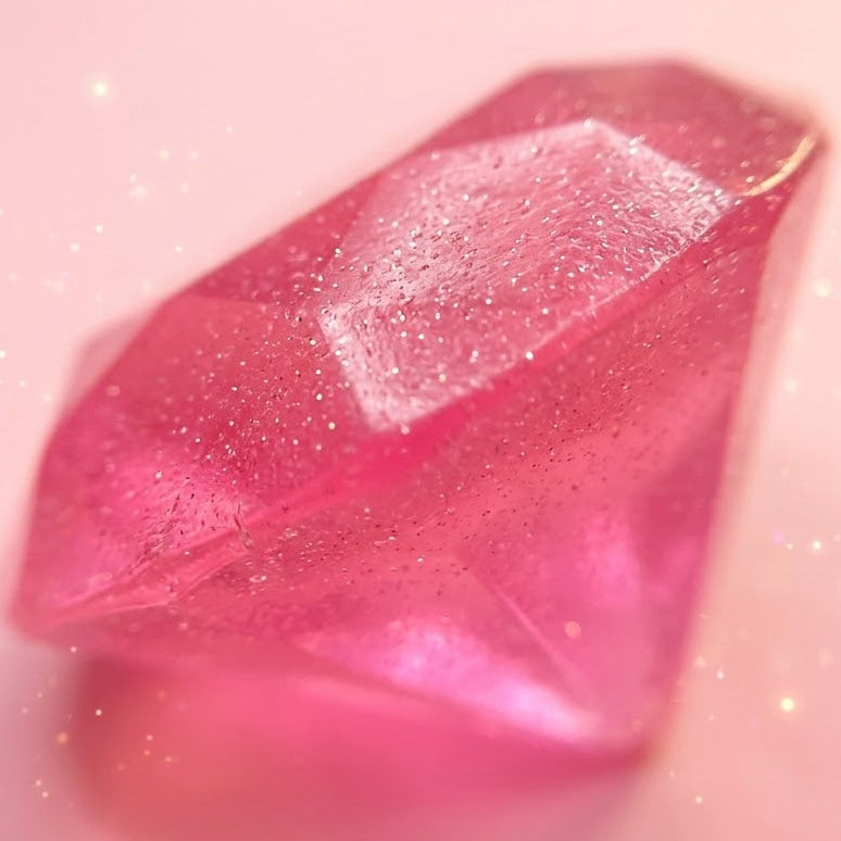 A faceted pink gemstone-like decorative piece with embedded glitter, photographed on a soft pink background with a dreamy, sparkly effect.