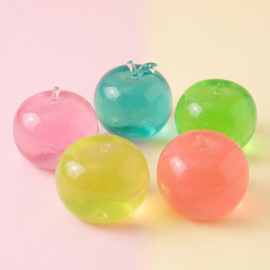 Five translucent, apple-shaped decorative objects in pink, teal, green, yellow, and coral orange, arranged on a soft pastel pink and cream background.
