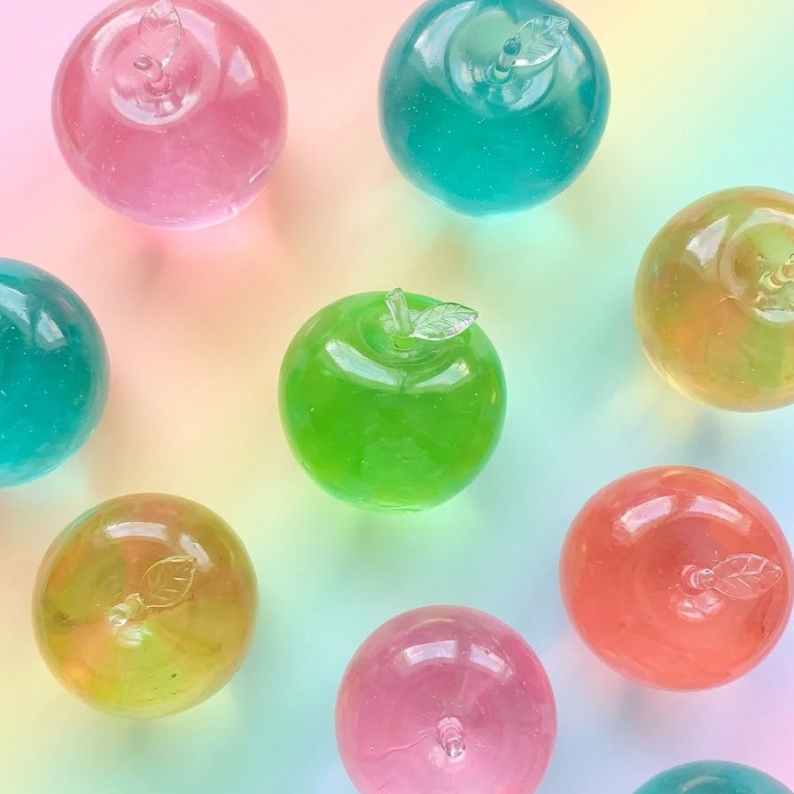 A set of translucent, apple-shaped resin pendants or charms in various pastel colors including pink, green, blue, orange, and yellow, arranged on a soft pastel rainbow gradient background.