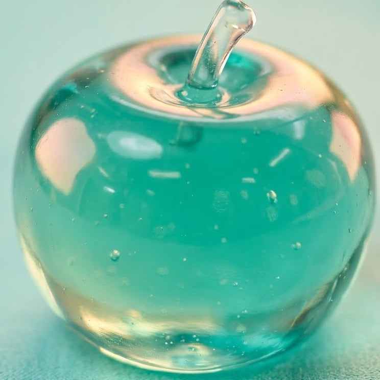 A small translucent glass apple-shaped object with a stem, filled with aqua blue liquid or gel, photographed on a soft light blue background.