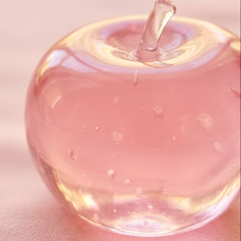 A translucent pink glass apple-shaped object with a small stem, photographed close up on a soft pink surface.