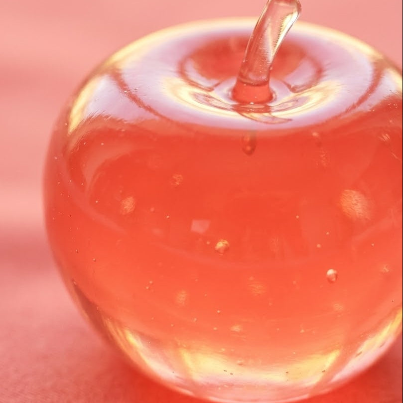 A close-up of a translucent, apple-shaped glass vessel filled with pink liquid, set against a matching pink background.