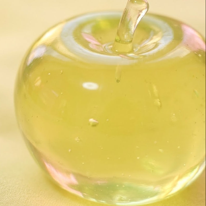 A translucent yellow-green apple-shaped glass object with a small stem, photographed close up on a soft yellow background.