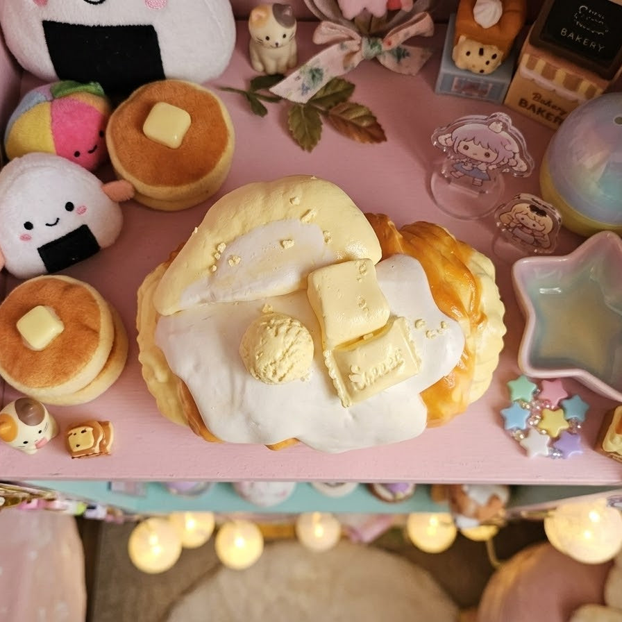 A cute, pastel-themed tabletop display featuring a central fluffy pancake with butter and cream, surrounded by plush toys, small pastries, stickers, and decorative items on a pink background.