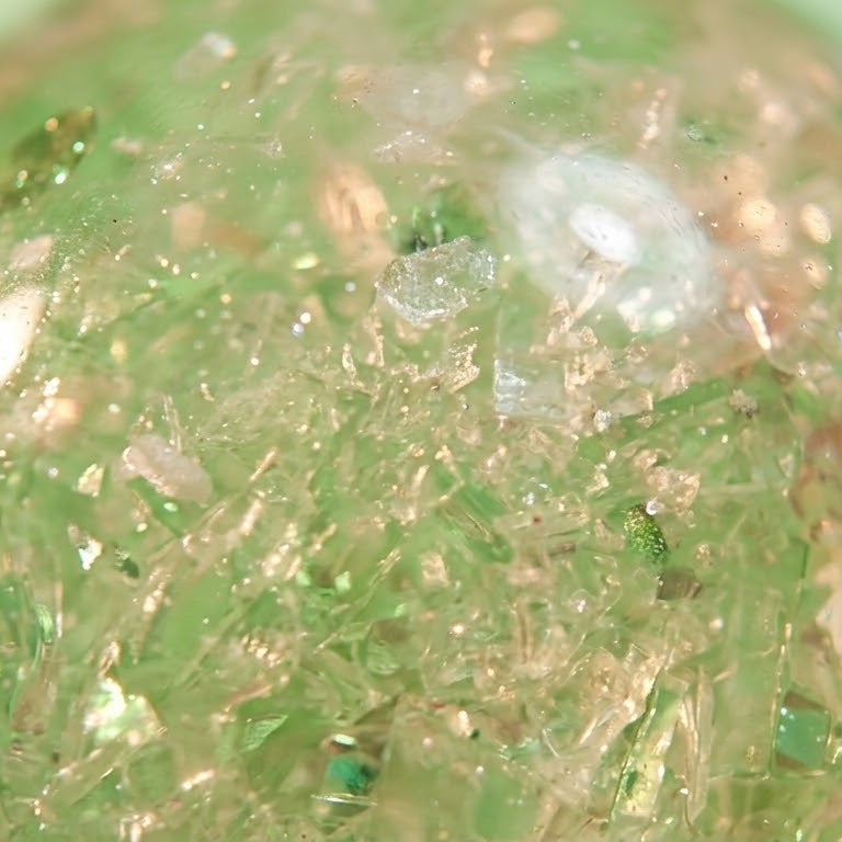 A close-up of a translucent green decorative sphere filled with glitter, crystals, and small reflective flecks on a pale green background.