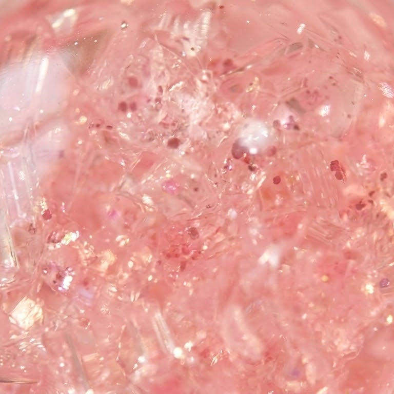 A close-up of a translucent pink sphere filled with pink glitter, clear crystal-like fragments, and iridescent flecks on a soft pink background.