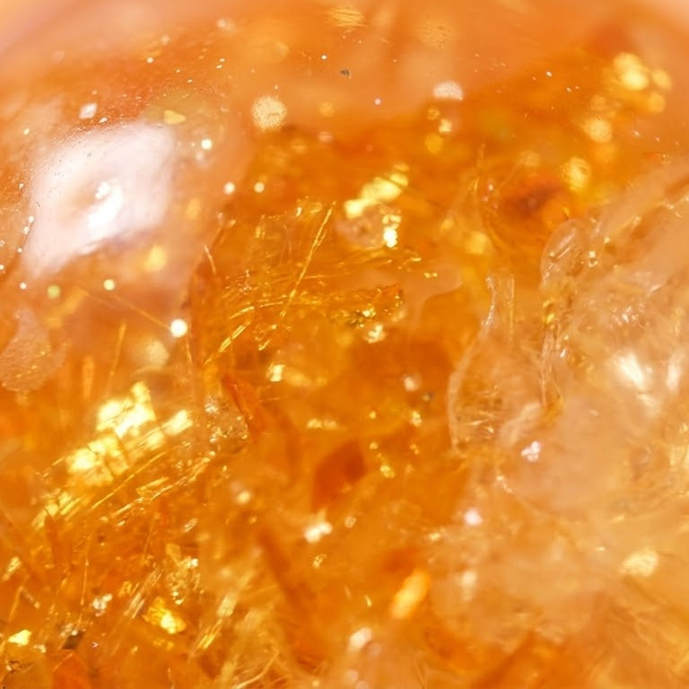 A close-up of a translucent amber-orange sphere filled with shimmering gold glitter and crystalline texture, set against a soft peach background.