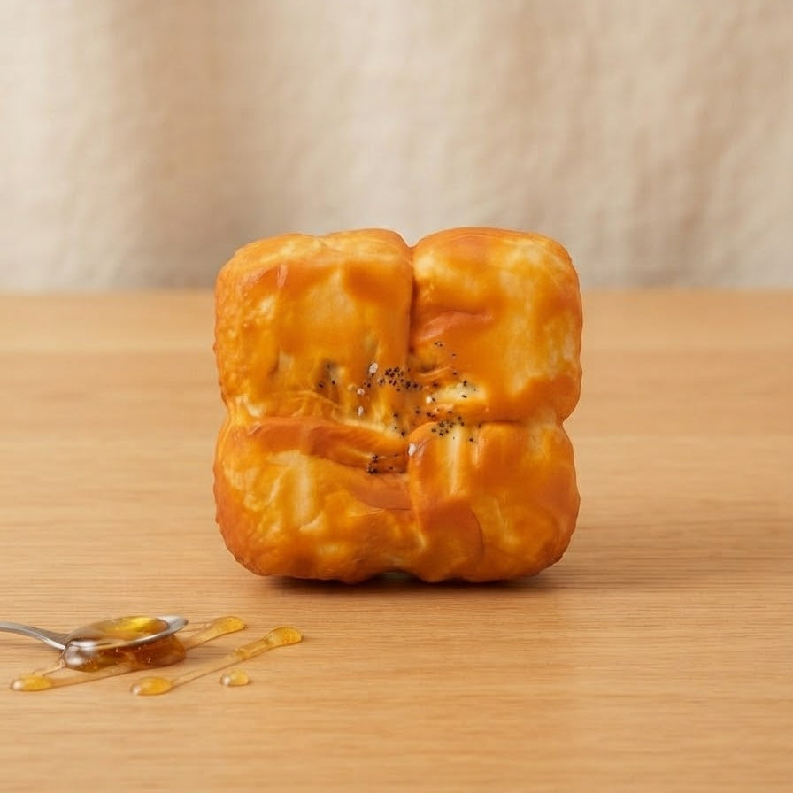 A golden-brown baked pastry or bread piece with a glossy orange glaze, standing upright on a light wood surface with a small spoon of honey nearby against a soft beige background.