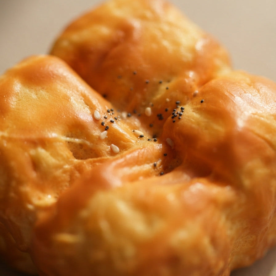 A close-up shot of a golden-brown baked bread roll, possibly a brioche or challah, topped with sesame seeds and poppy seeds, on a neutral beige background.
