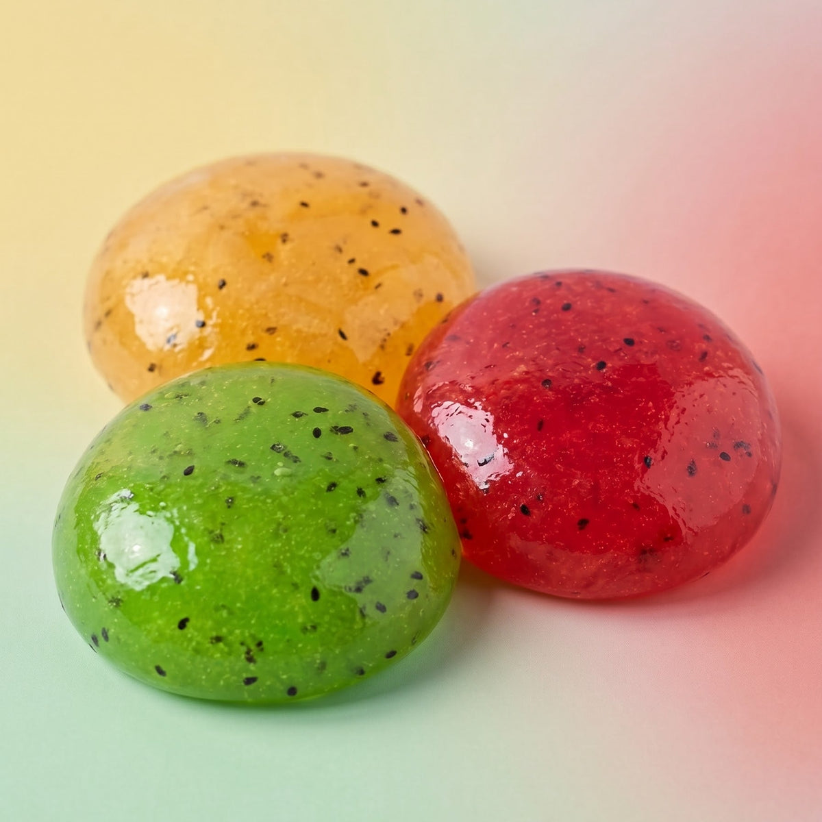 Three glossy, translucent gel candies in green, orange, and red, each speckled with tiny black seeds, arranged on a pastel gradient background.
