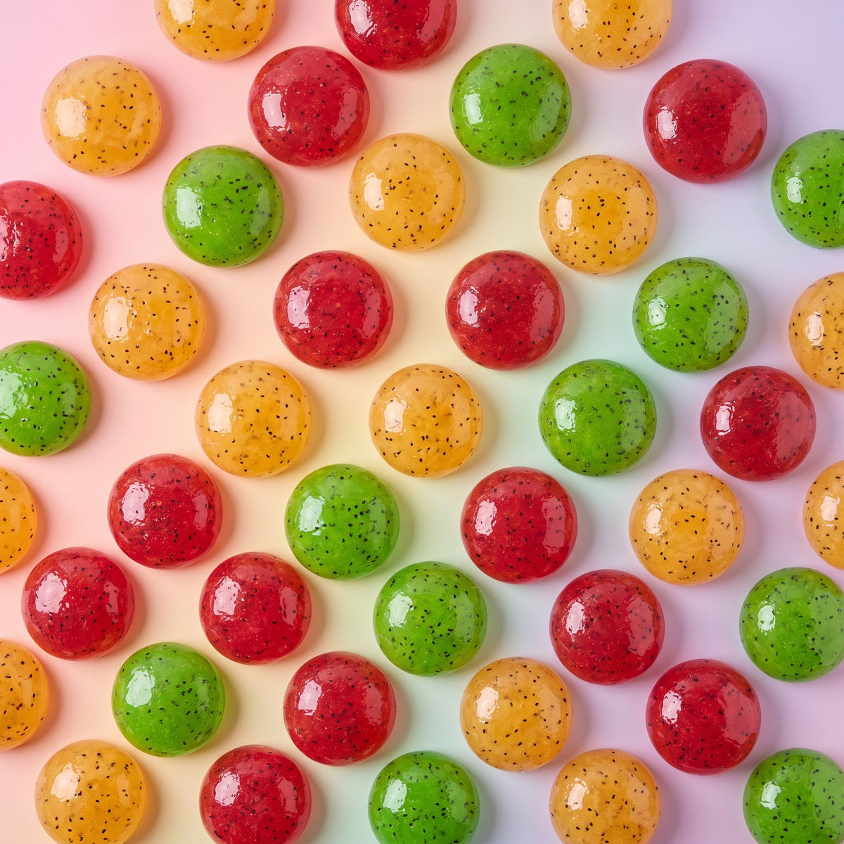 A close-up overhead arrangement of glossy round candies in red, green, and translucent orange-yellow, with tiny dark speckles, set against a pale pink background.