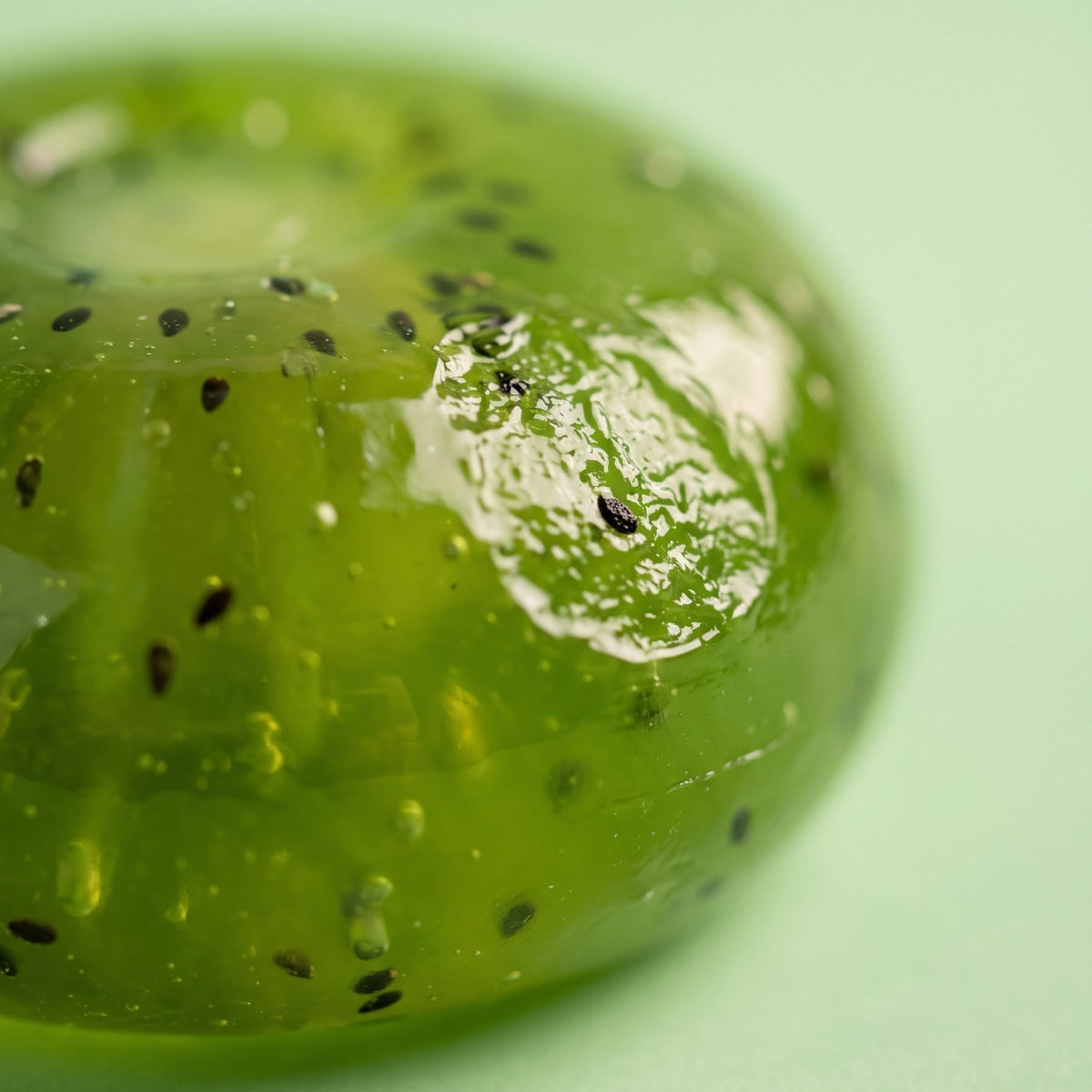 A close-up of a kiwi-shaped gel or candy with green flesh, black seeds, and a glossy finish on a light green background.