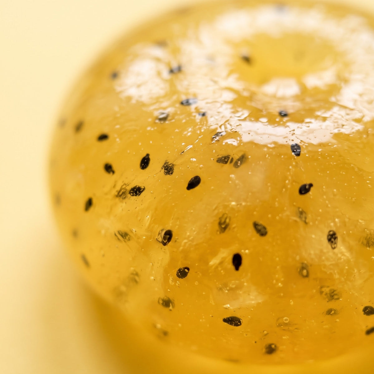 A close-up of a translucent yellow gelatin or jelly dessert sphere with small black seed-like specks, photographed on a pale yellow background.