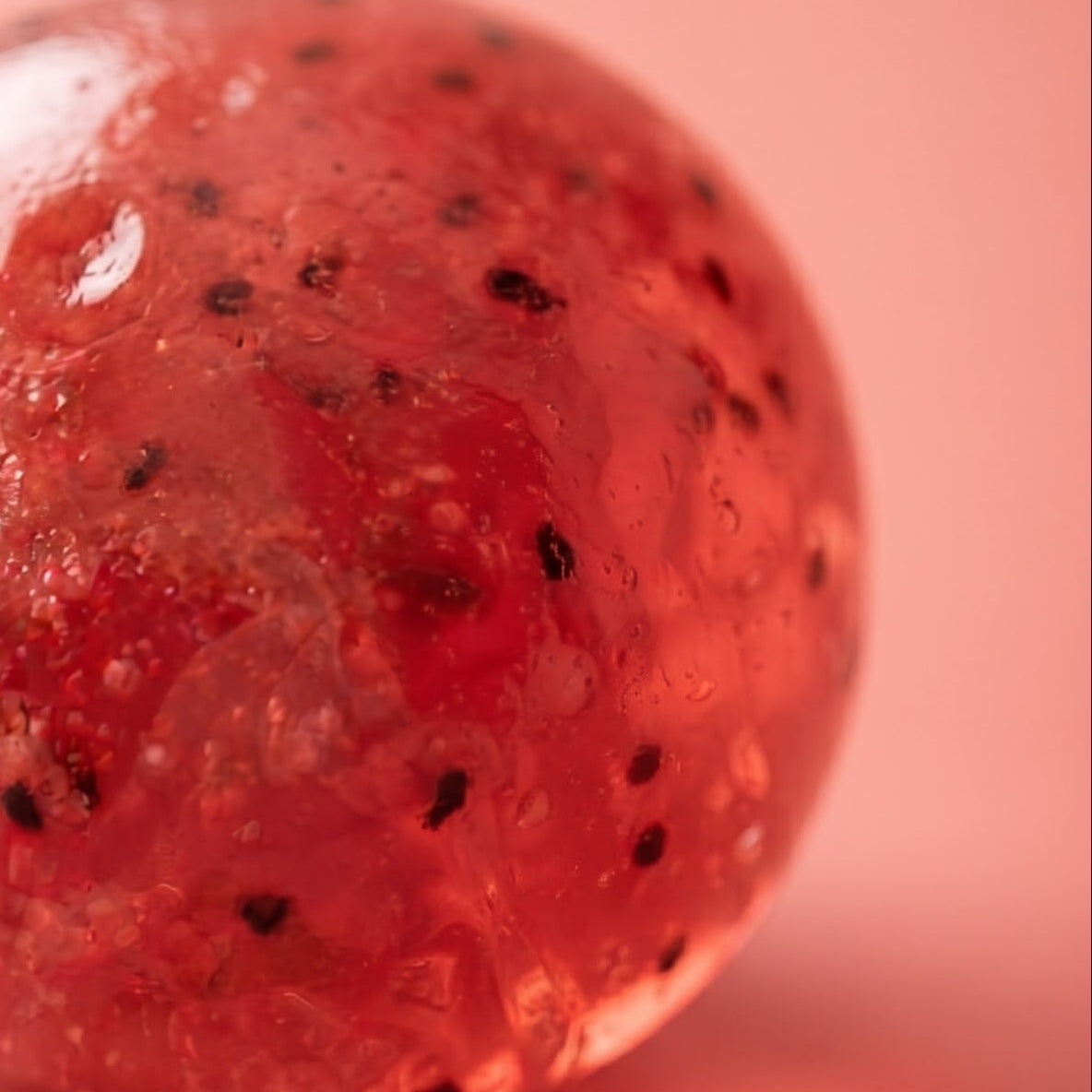 A translucent pinkish-red spherical object with small black speckles, photographed on a soft pink background.