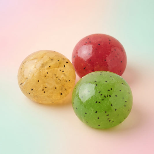 Three smooth, glossy stress balls in yellow, red, and green, each speckled with tiny black seeds, arranged on a pastel gradient background.