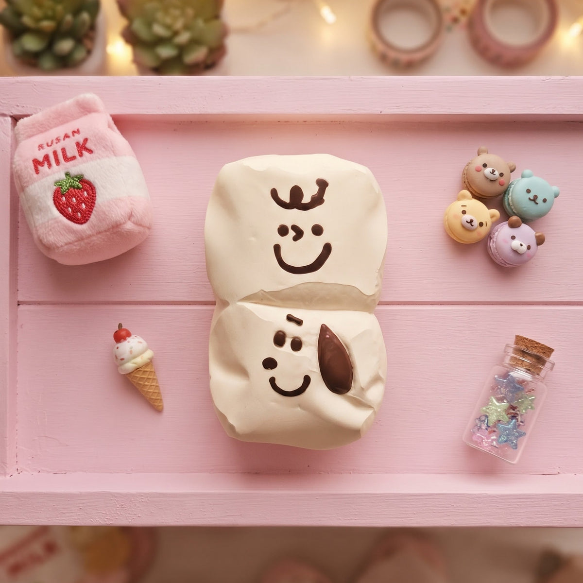 A styled flat-lay of a cream-colored squishy toy shaped like a split marshmallow with a smiling face, displayed on a pink wooden tray alongside a pink milk carton plush, a miniature ice cream cone, small character charms, and a tiny glitter bottle.