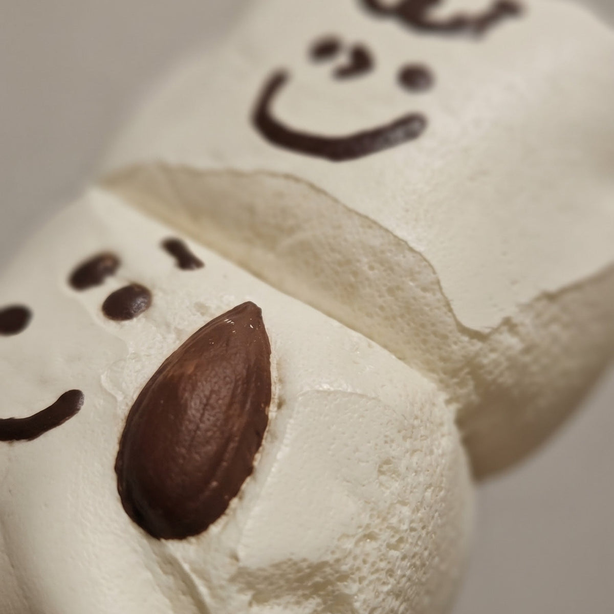 A close-up of a white marshmallow snowman decoration with a brown almond for a nose and simple dark facial features on a neutral background.