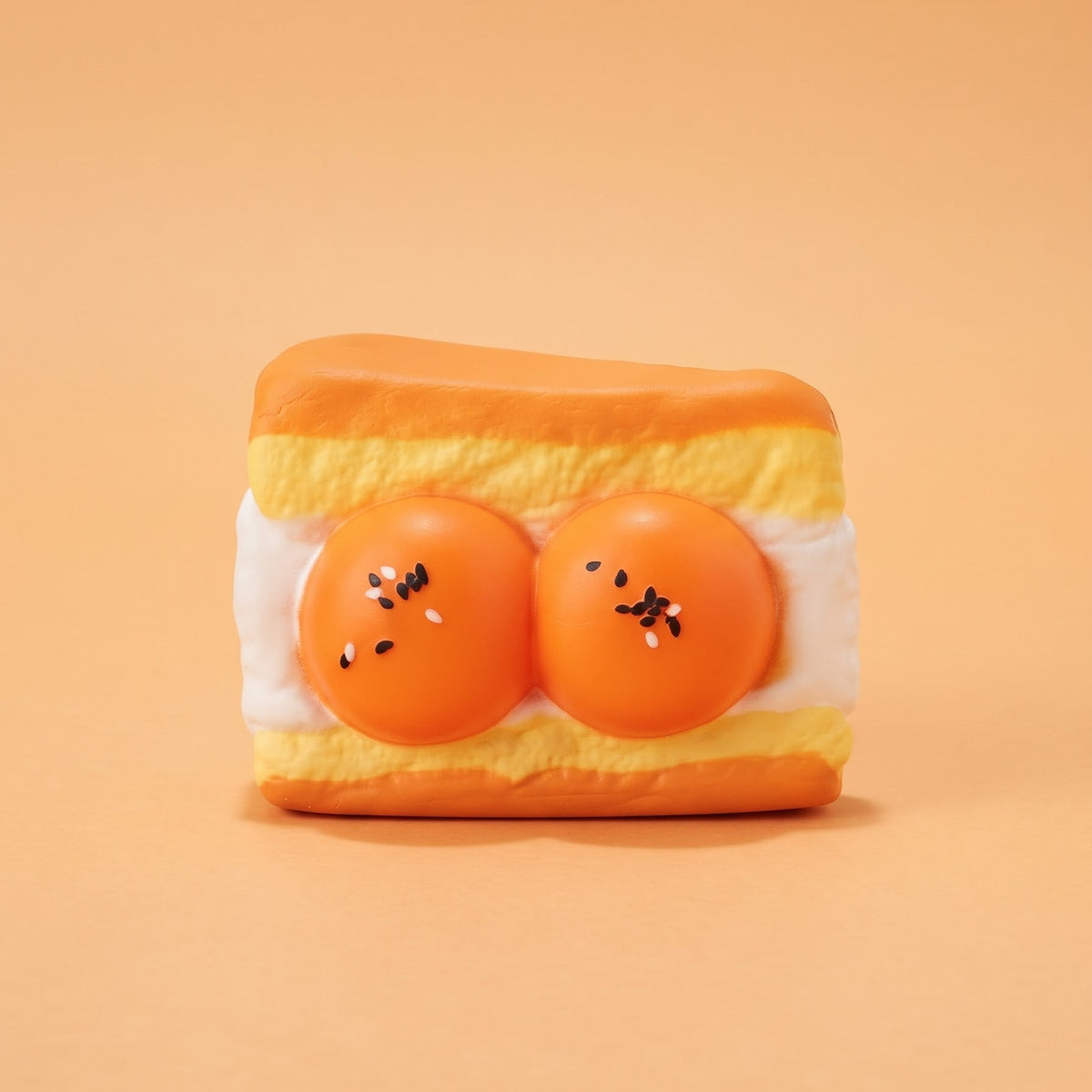 A novelty squishy toy shaped like a toasted sandwich with two orange egg yolks on the front, set against a solid peach-colored background.