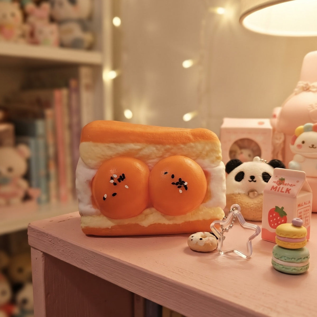 A cute, sandwich-shaped squishy toy with two orange eggs and black spider details, displayed on a pink shelf next to other small plush toys and accessories in a cozy room setting.