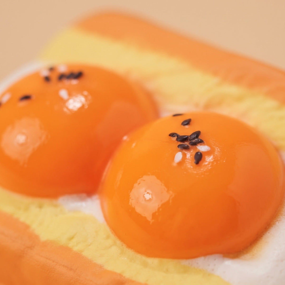 A close-up of a novelty food item shaped like a breakfast sandwich, featuring two glossy orange egg yolks on a yellow and white layered bun, with small black sesame seeds on top.