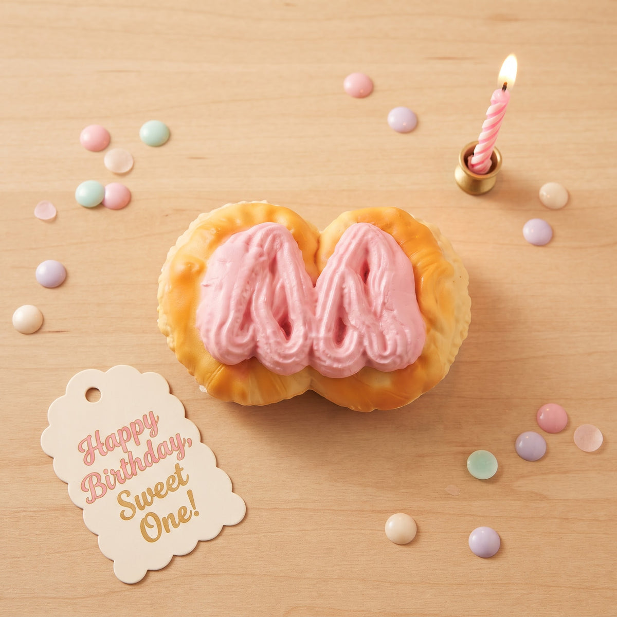 A heart-shaped pastry with pink frosting sits on a light wood surface, styled with pastel candy pieces, a small lit candle, and a paper tag that reads 'Happy Birthday, Sweet One!'.