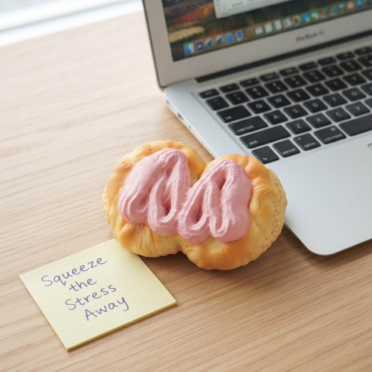 A novelty stress toy shaped like a pastry with pink frosting sits on a light wood desk next to a sticky note that reads 'Squeeze the Stress Away' and a partially visible laptop.