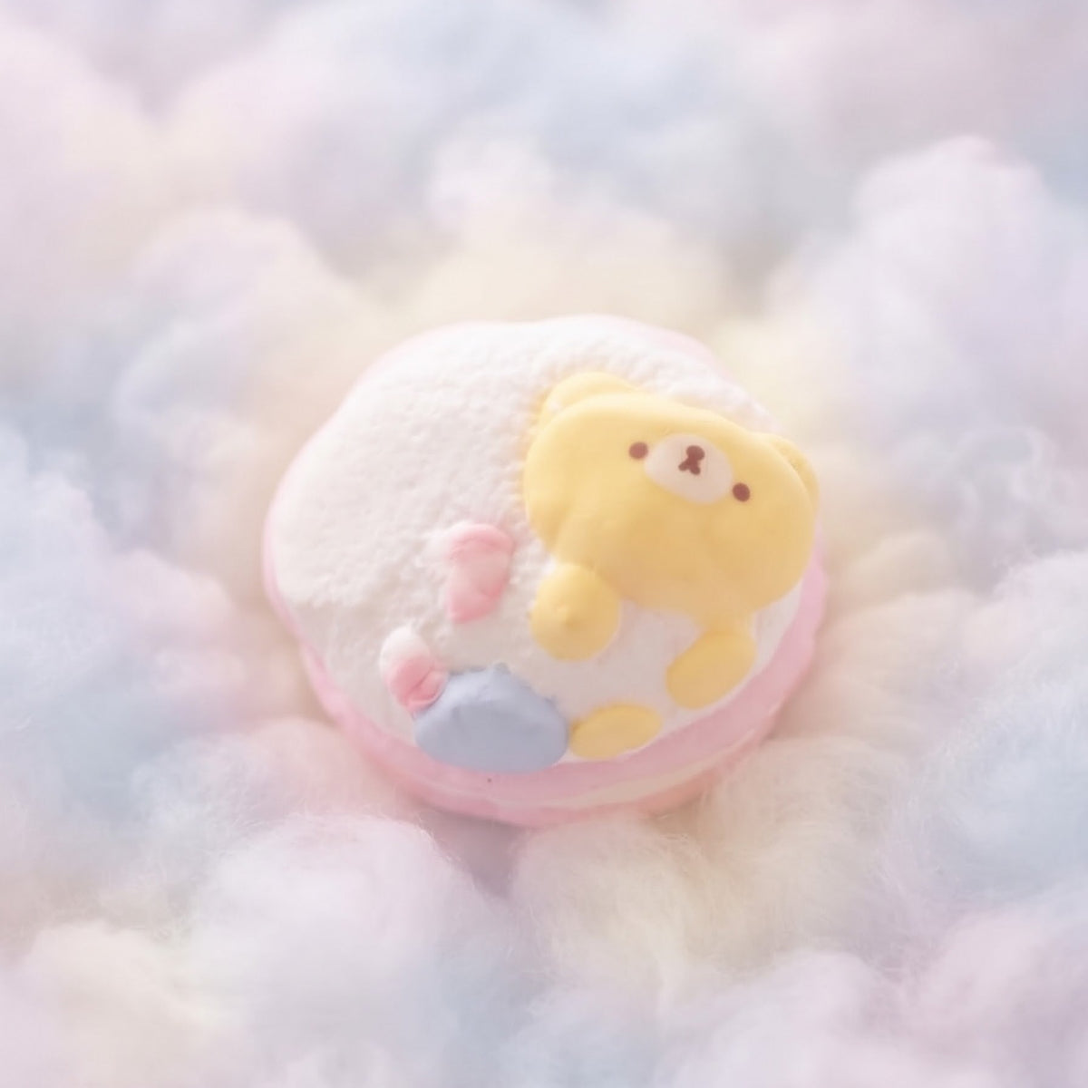A pastel macaron with a cute yellow bear face and small pink and blue details, resting on a bed of fluffy white, pink, and blue cotton candy.