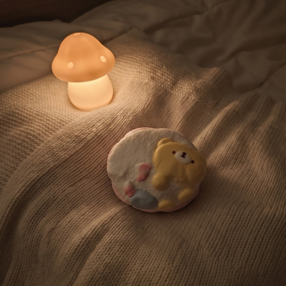 A cozy bedroom scene with a small mushroom-shaped glowing night light and a round plush bear toy resting on rumpled beige bedding.