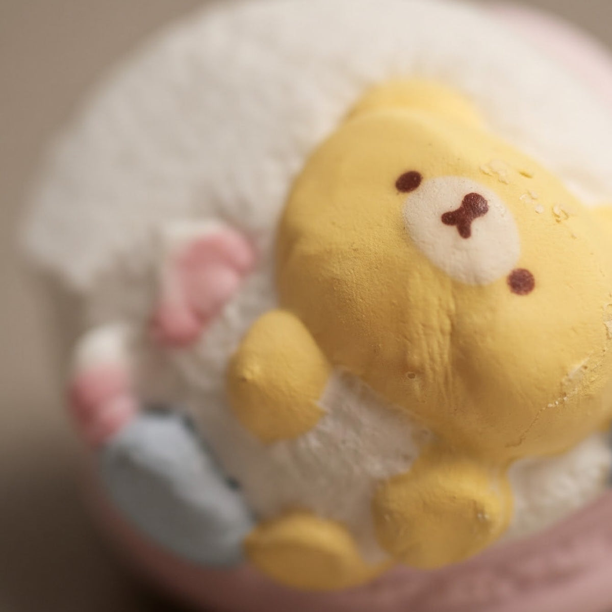 A close-up of a pastel macaron with a yellow bear face on top, nestled in white frosting, with pink and blue accents, set against a soft beige background.